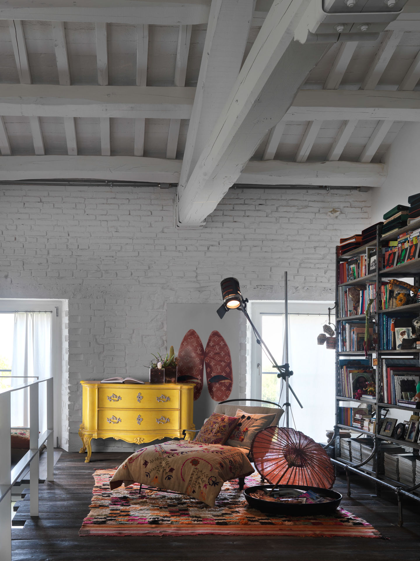 Living in mansarda con travi bianche, comò classico bombato giallo acceso, chaise longue con tessuti etnici e libreria industriale in ferro. Progetto di Bertelè Mobili (Verona)