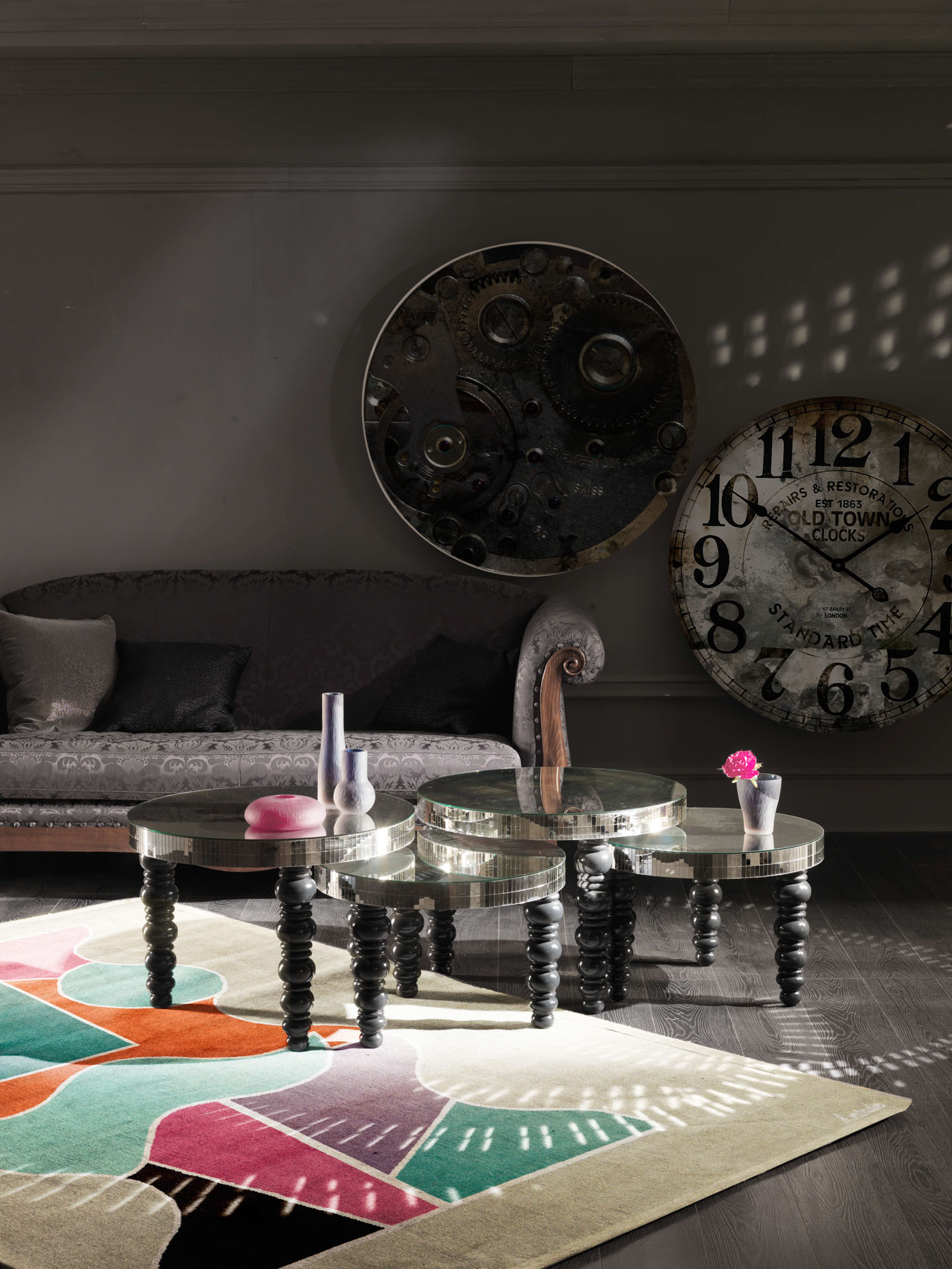 Set di quattro tavolini rotondi specchiati con gambe tornite nere, tappeto geometrico colorato e orologi vintage giganti sullo sfondo. Progetto di interior design di Bertelè Mobili (Verona)